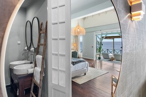 A modern bedroom with a large bed, a bathroom with a sink and mirror, and a view of the ocean through an open door.