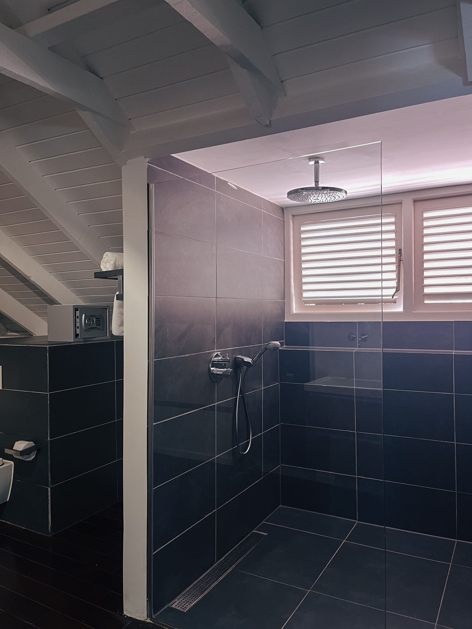 A modern bathroom with dark tiles, a shower, a window with blinds, and a ceiling with exposed beams.