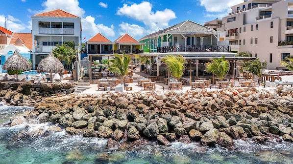 A beachfront hotel with outdoor seating, a rocky shoreline, and buildings with red-tiled roofs. - BijBlauw Hotel (Willemstad)