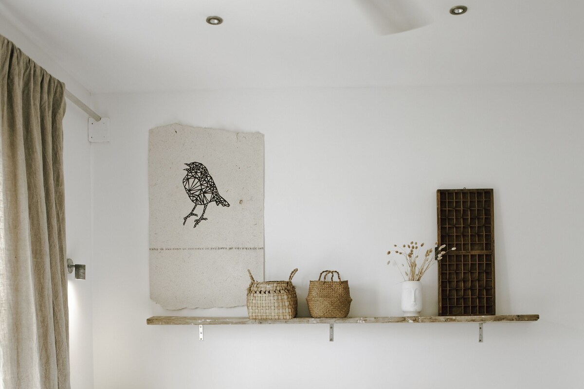A bedroom with a bed, two bedside tables, a hanging shelf with baskets and a vase, and a wall-mounted artwork of a bird.