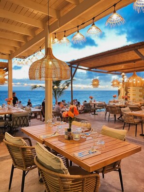An outdoor dining area with wooden tables and wicker chairs, overlooking the sea. - BijBlauw Hotel (Willemstad)