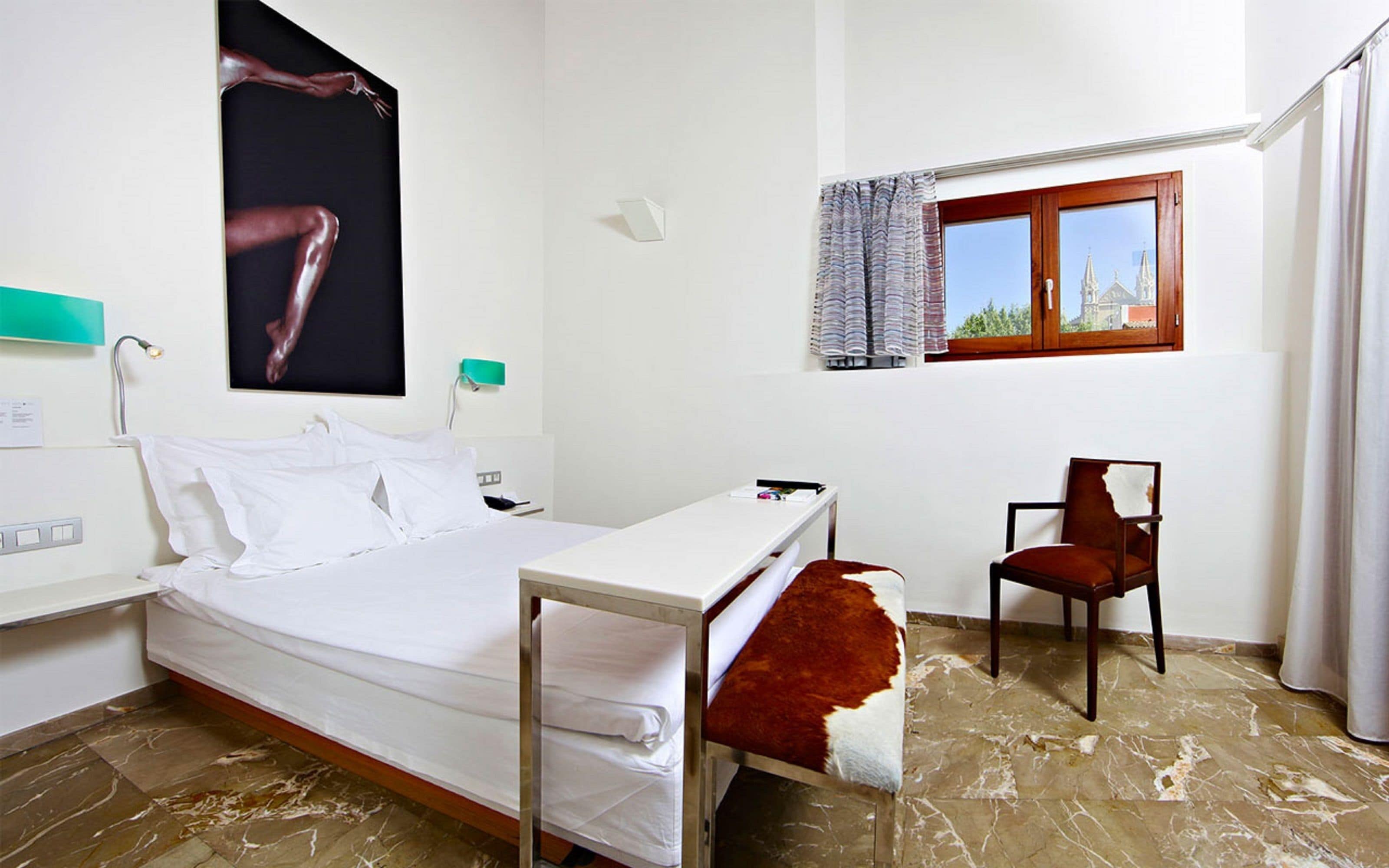 A hotel room with a bed, a desk, a chair, a window with curtains, and a painting on the wall.