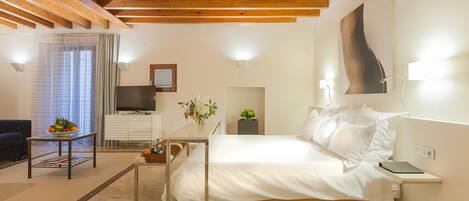 Un dormitorio moderno con techo de madera, cama con ropa blanca, mesita de noche con un libro, una planta pequeña y un televisor en un aparador blanco.