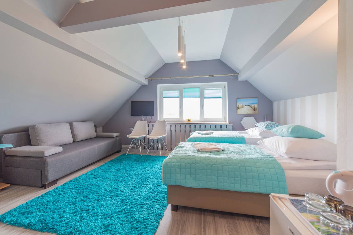 Un dormitorio moderno con techo inclinado, un ventanal grande, un sofá, una cama y un tapete color turquesa.