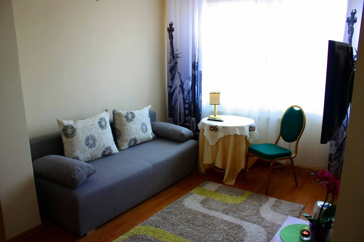 Una sala de estar con un sofá gris, una mesita redonda con una lámpara, una silla verde y un televisor.