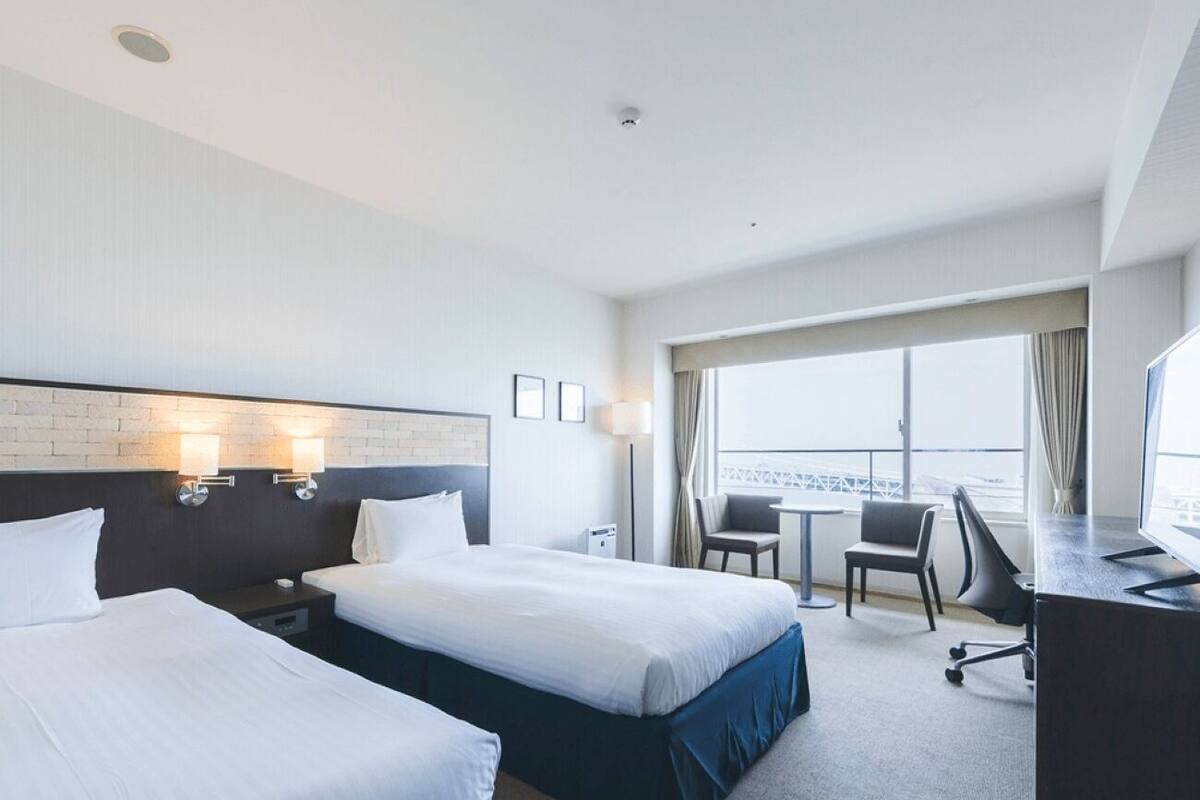 A hotel room with two beds, a desk, a chair, and a large window offering a view.