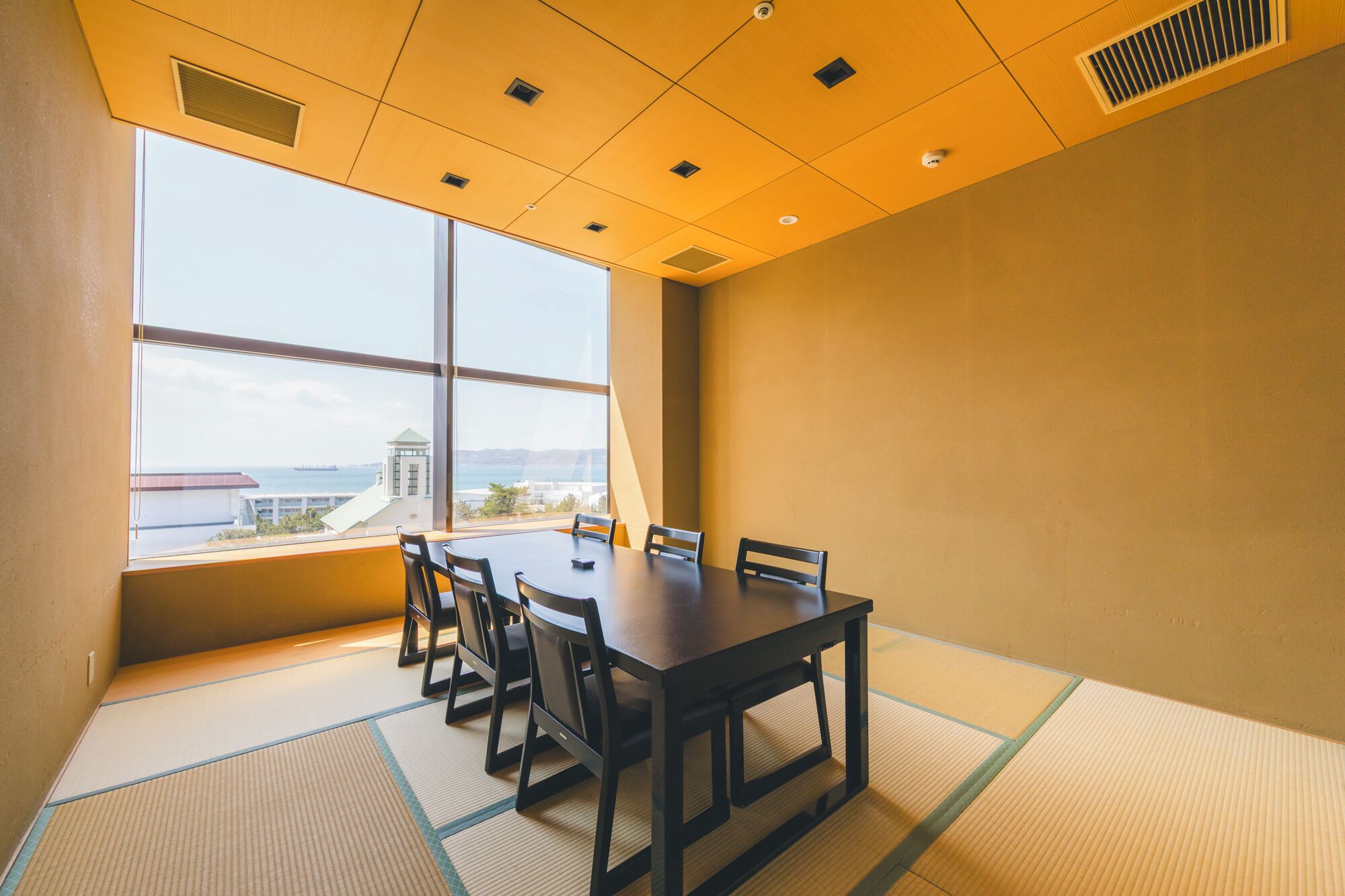 A conference room with a large table and chairs, a view of the sea through large windows, and a high ceiling with recessed lighting.