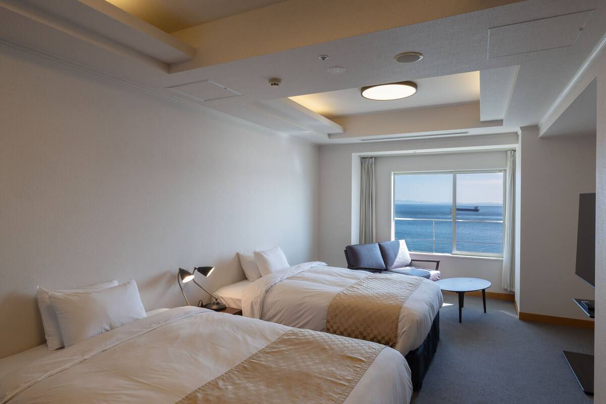 A hotel room with two beds, a desk, a chair, a TV, and a large window with a view of the sea.