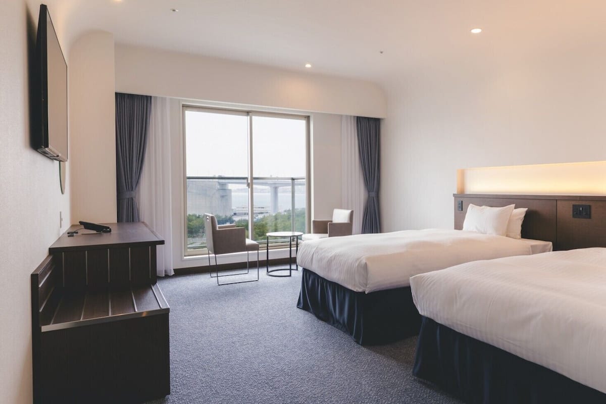 A hotel room with two beds, a desk, a chair, and a large window with curtains.