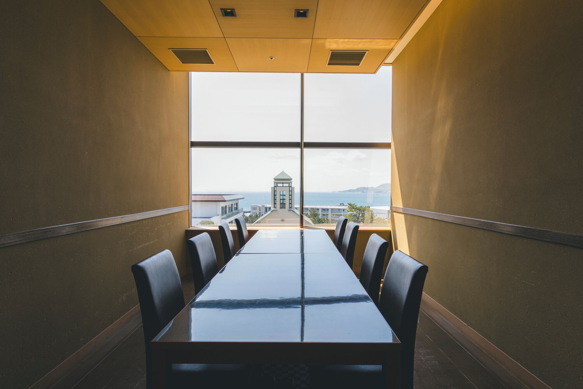 A conference room with a long table and chairs, large windows offering a view of a building and the sea.