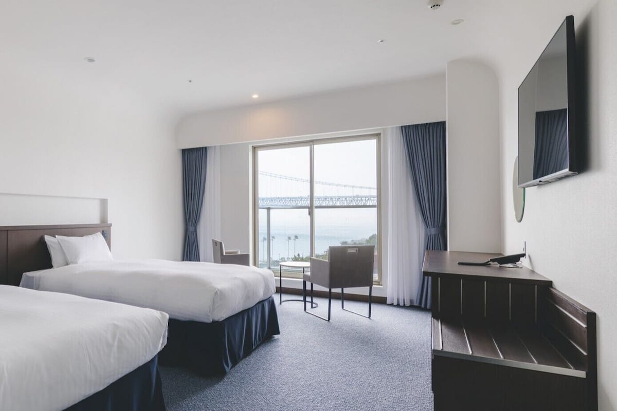 A hotel room with two beds, a desk, a chair, and a large window with a view of a bridge.