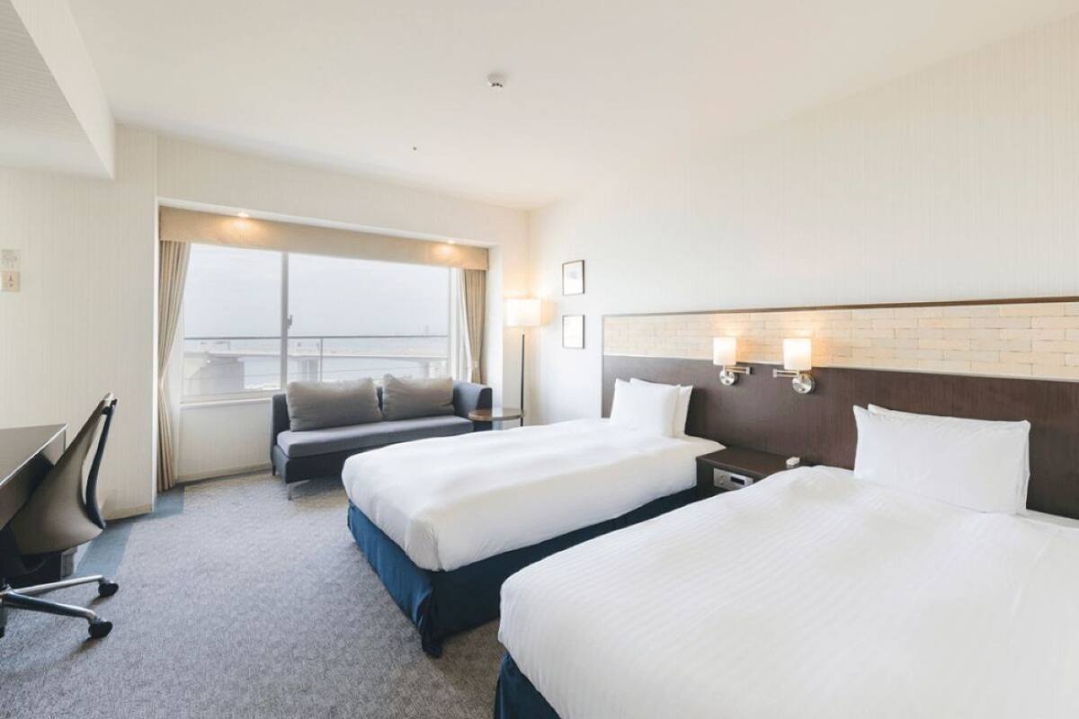 A hotel room with two beds, a desk, a chair, and a large window.
