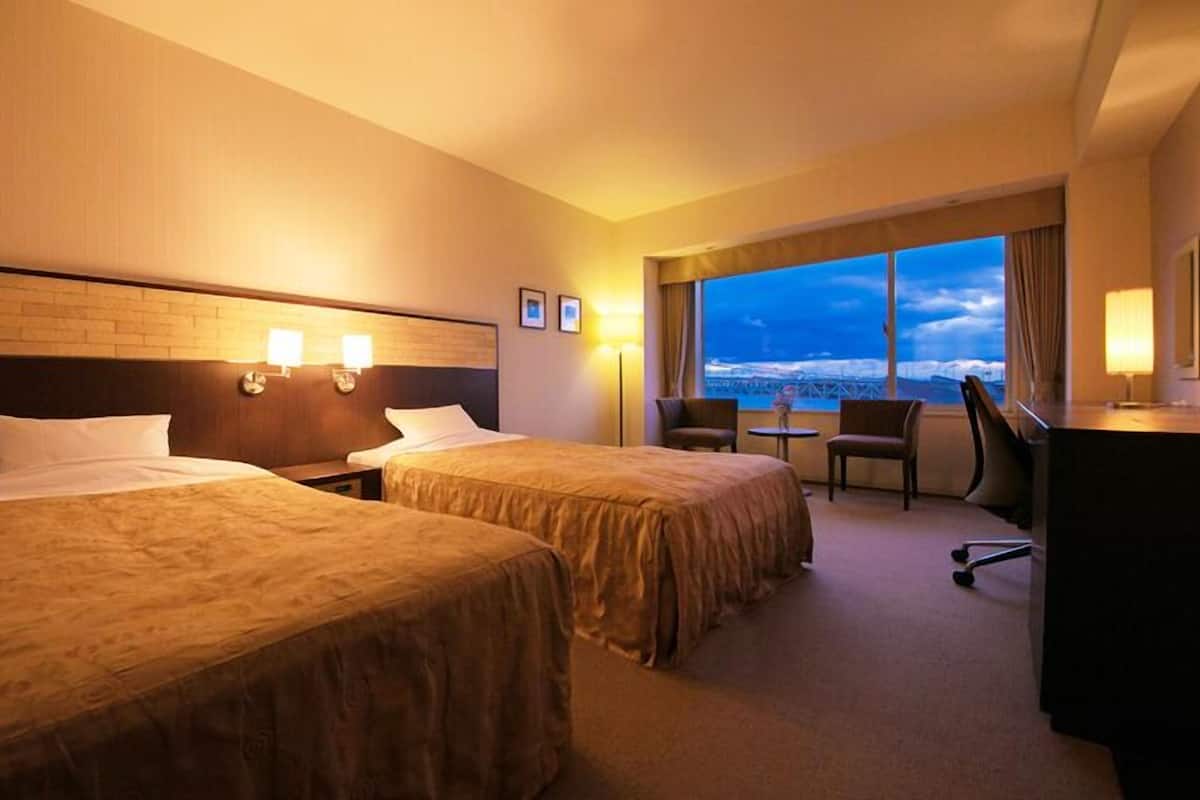 A hotel room with two beds, a desk, a chair, and a large window with a view.