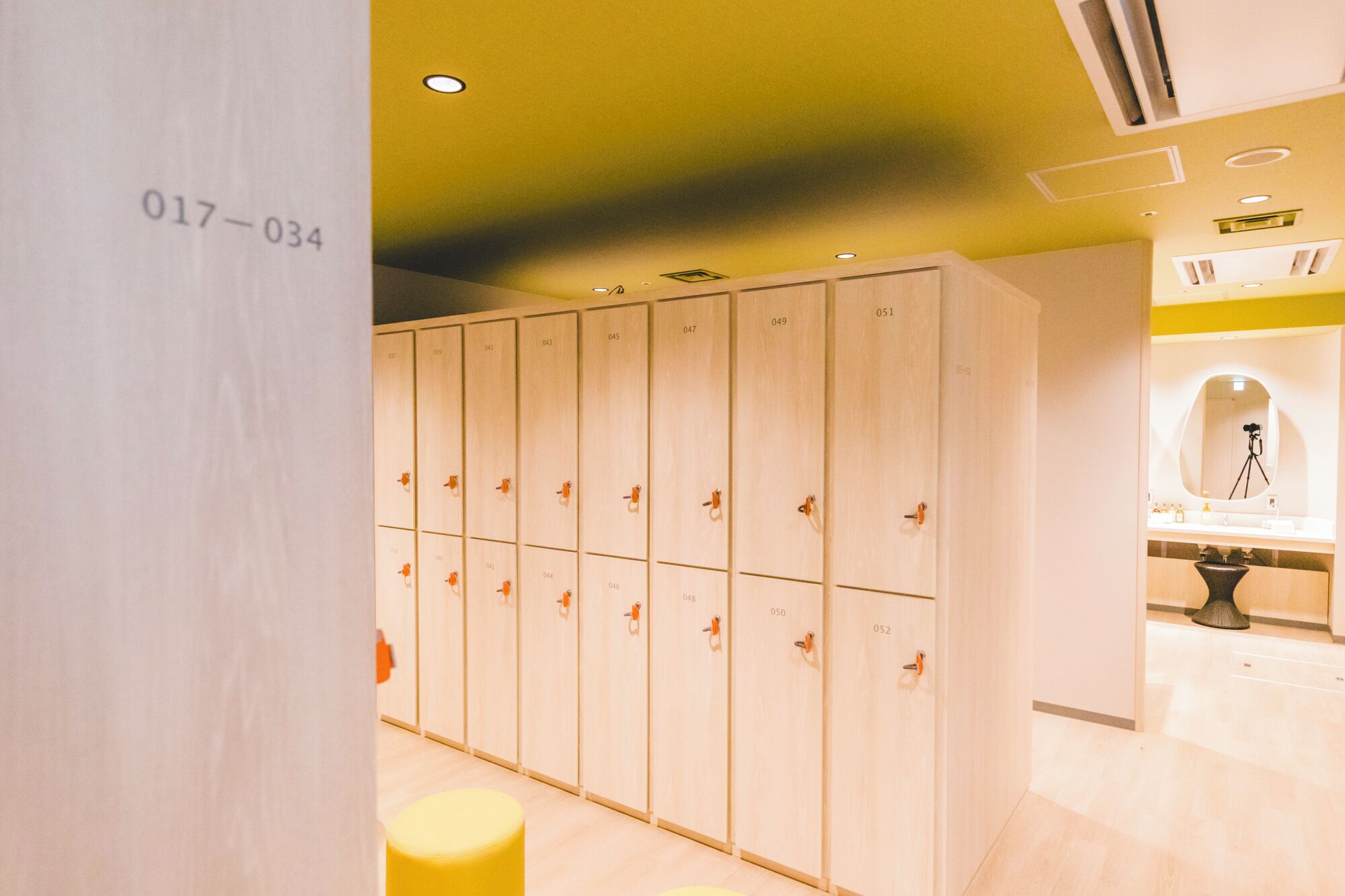 A row of wooden lockers with numbers, a yellow stool, and a sink area with a mirror.