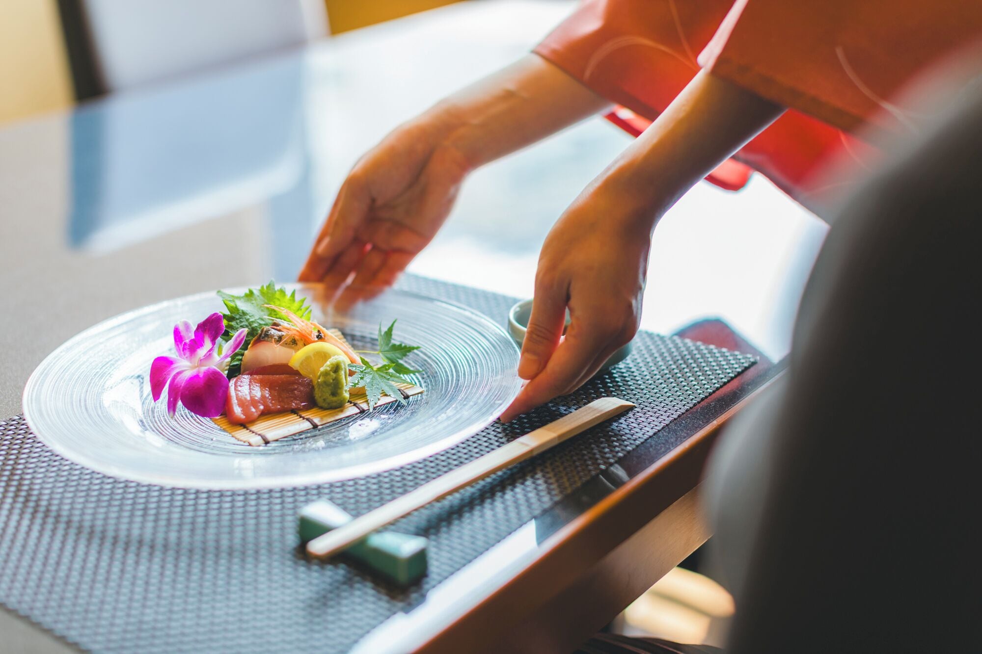 A person placing a dish on a clear glass plate with food and garnish on a textured mat.