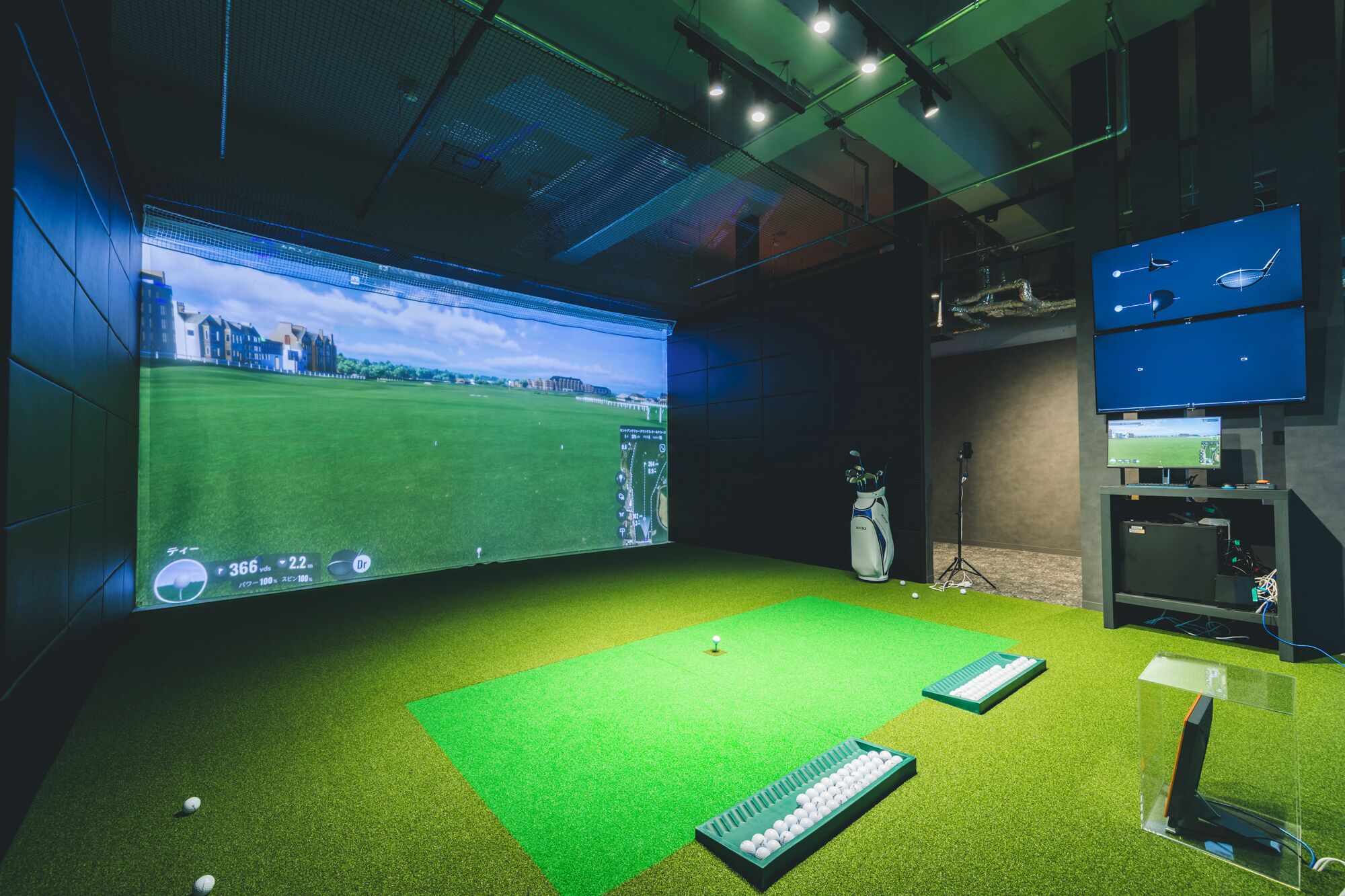 A virtual golf simulation room with a large screen displaying a golf course, a golf bag, a keyboard, and a golf ball on a green mat.