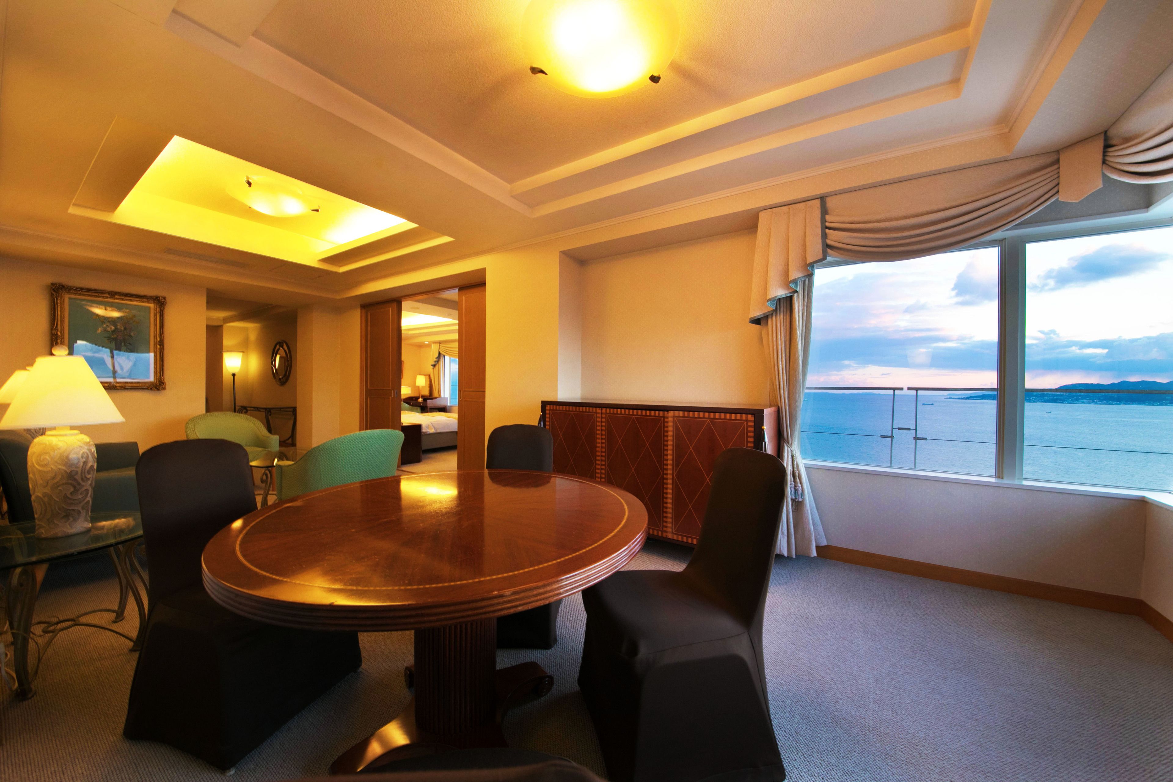 Une chambre d’hôtel avec une table ronde, quatre chaises, une lampe, un tableau et une vue sur la mer.