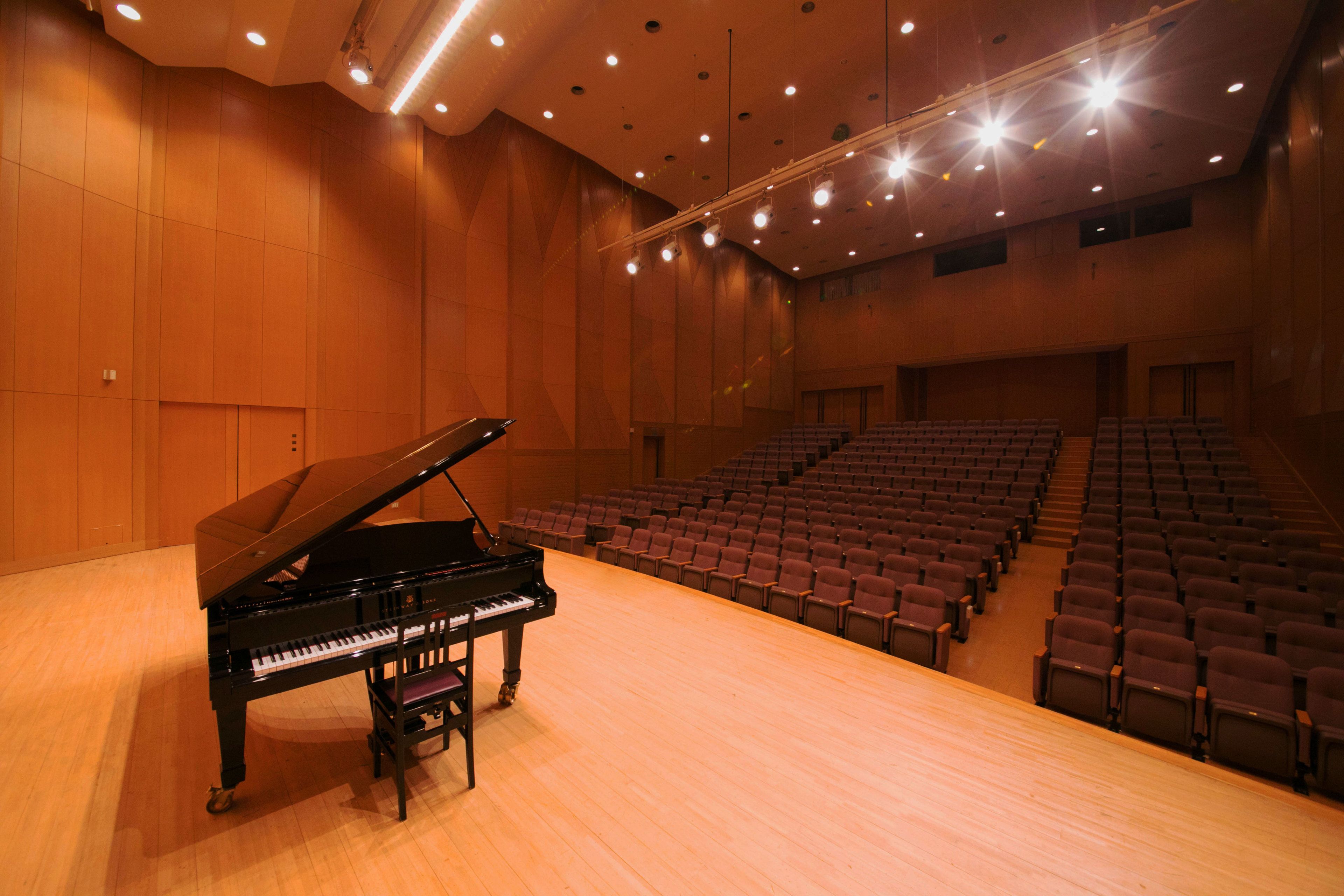 An auditorium with a grand piano, rows of seating, and a high ceiling with recessed lighting.