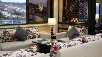 Un rincón acogedor con sofás y sillones de estampado floral, una mesita con lámpara, y una ventana que ofrece vistas a un pueblo.
