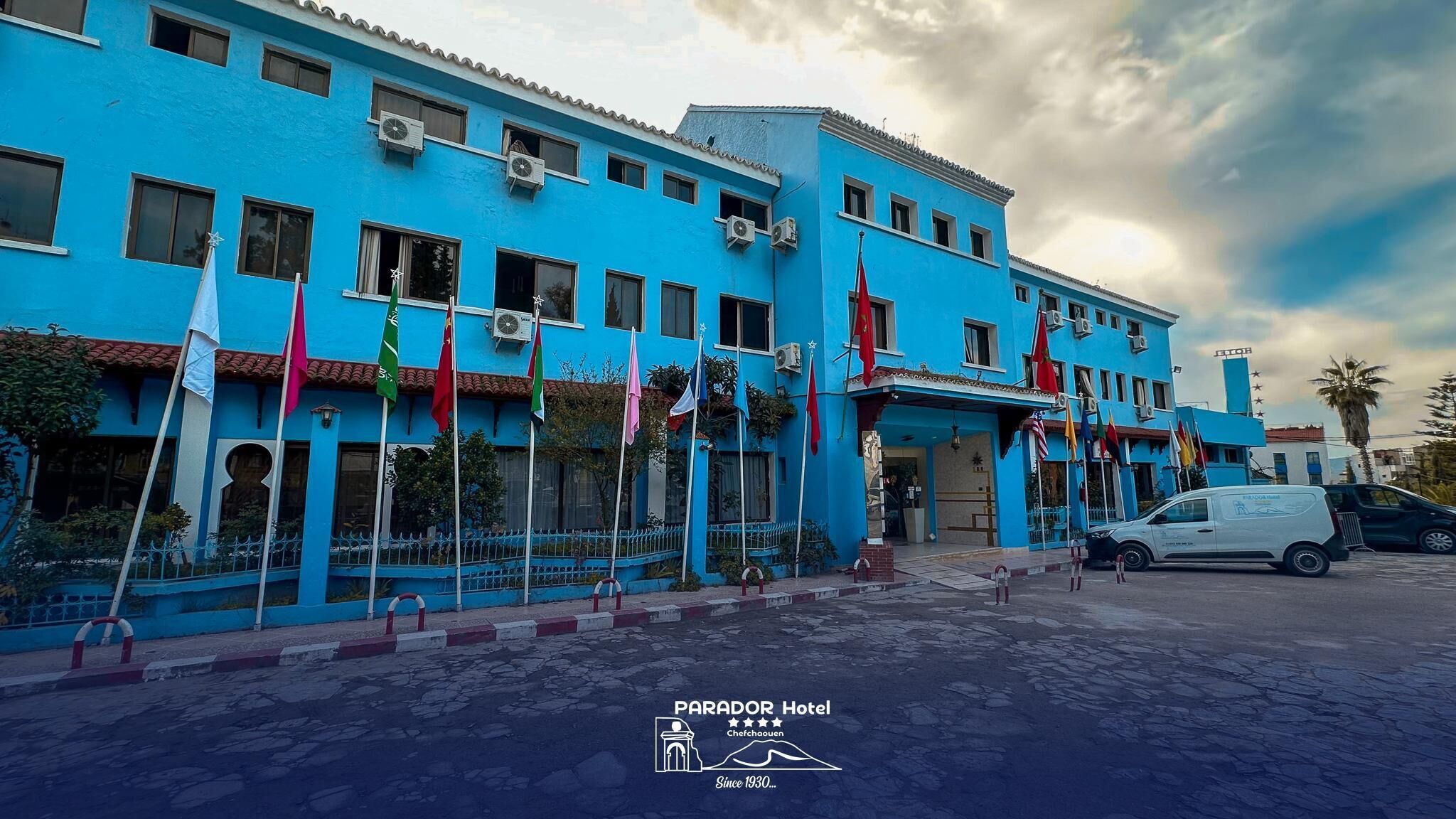 Un hotel con fachada azul, varias banderas y un estacionamiento.