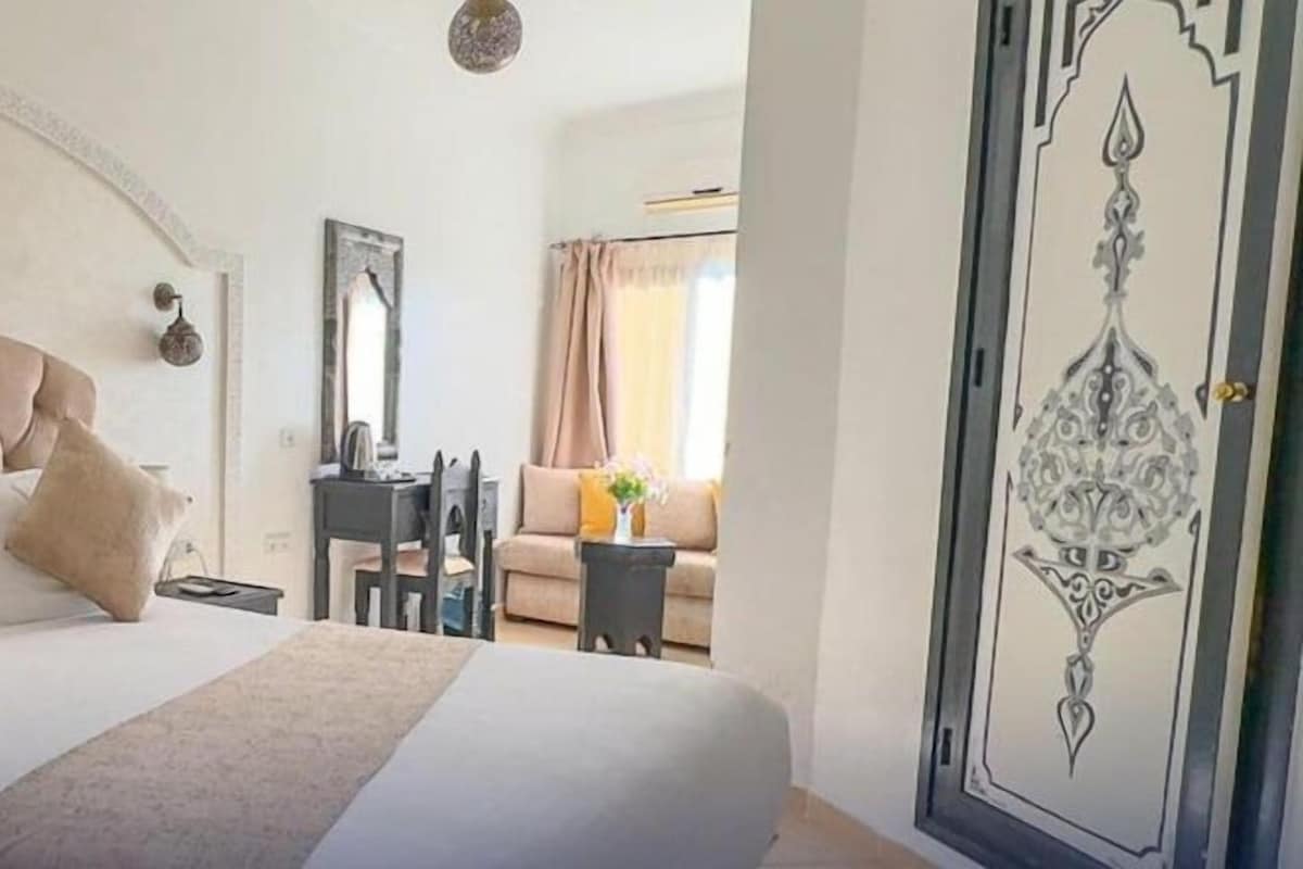 A hotel room with a large bed, a nightstand with a lamp, a chair, a small table, and a framed artwork on the wall.