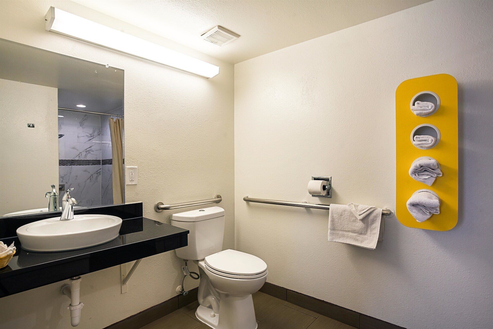 A bathroom with a sink, toilet, and a mirror. There is a towel rack with towels and a yellow towel holder with folded towels.