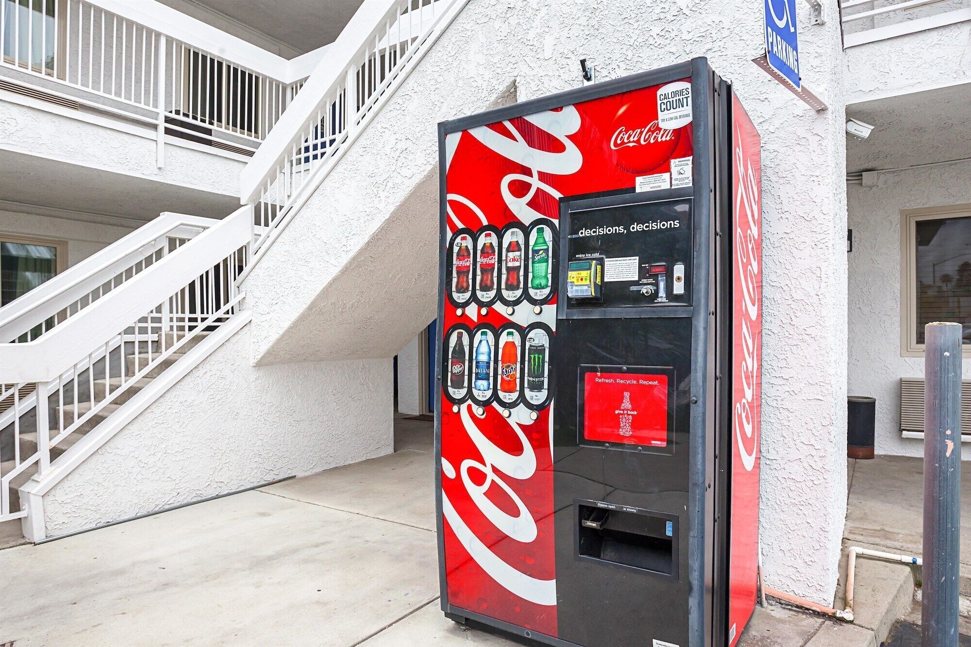 A Coca-Cola branded vending machine with various drink options, located outside a building with a white exterior and a staircase.