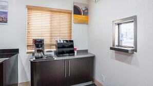 Une petite kitchenette équipée d’une machine à café, d’un distributeur d’eau et d’une cafetière. Il y a une fenêtre avec des stores, une pancarte au-dessus de la fenêtre et une petite fenêtre sur le côté droit.