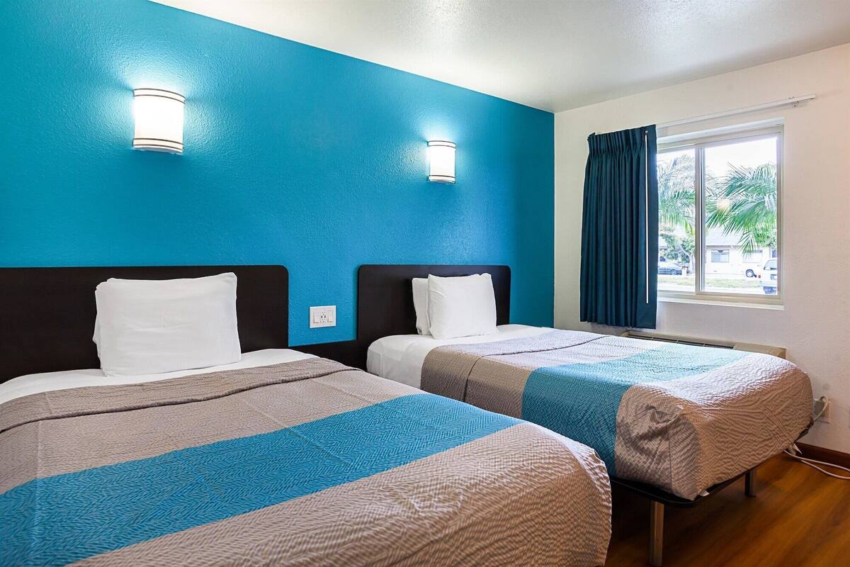 Ein Hotelzimmer mit zwei Betten, jeweils mit Kopfteil und weißen Kissen. Zwei Wandleuchten und ein Fenster mit Vorh ängen sind vorhanden.