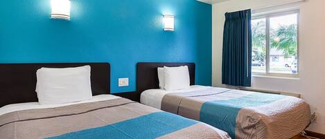 A hotel room with two beds, each with a headboard and white pillows. There are two wall-mounted light fixtures and a window with curtains.