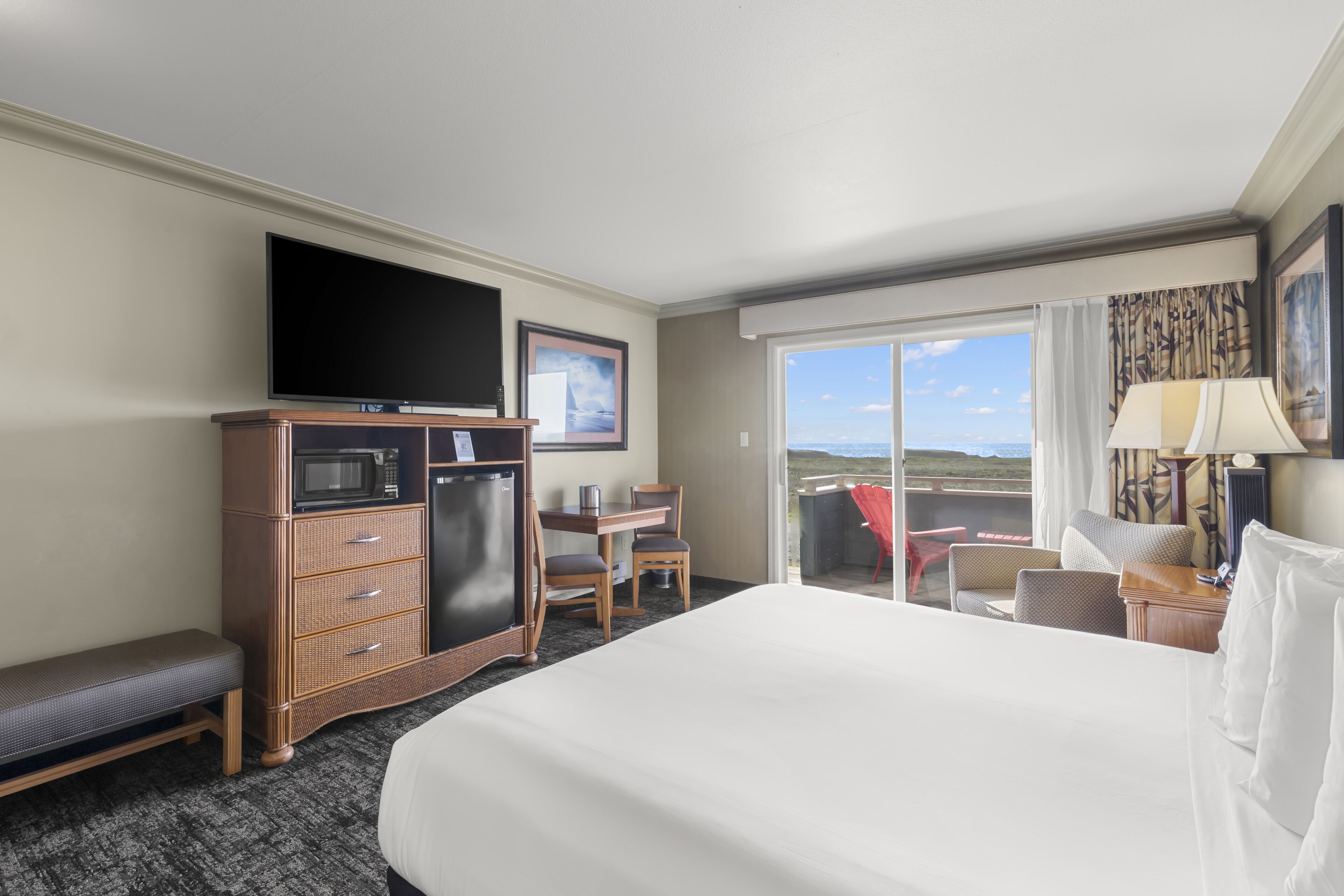 A hotel room with a large bed, a flat-screen TV, a minibar, a desk with a chair, and a view of the outdoors.