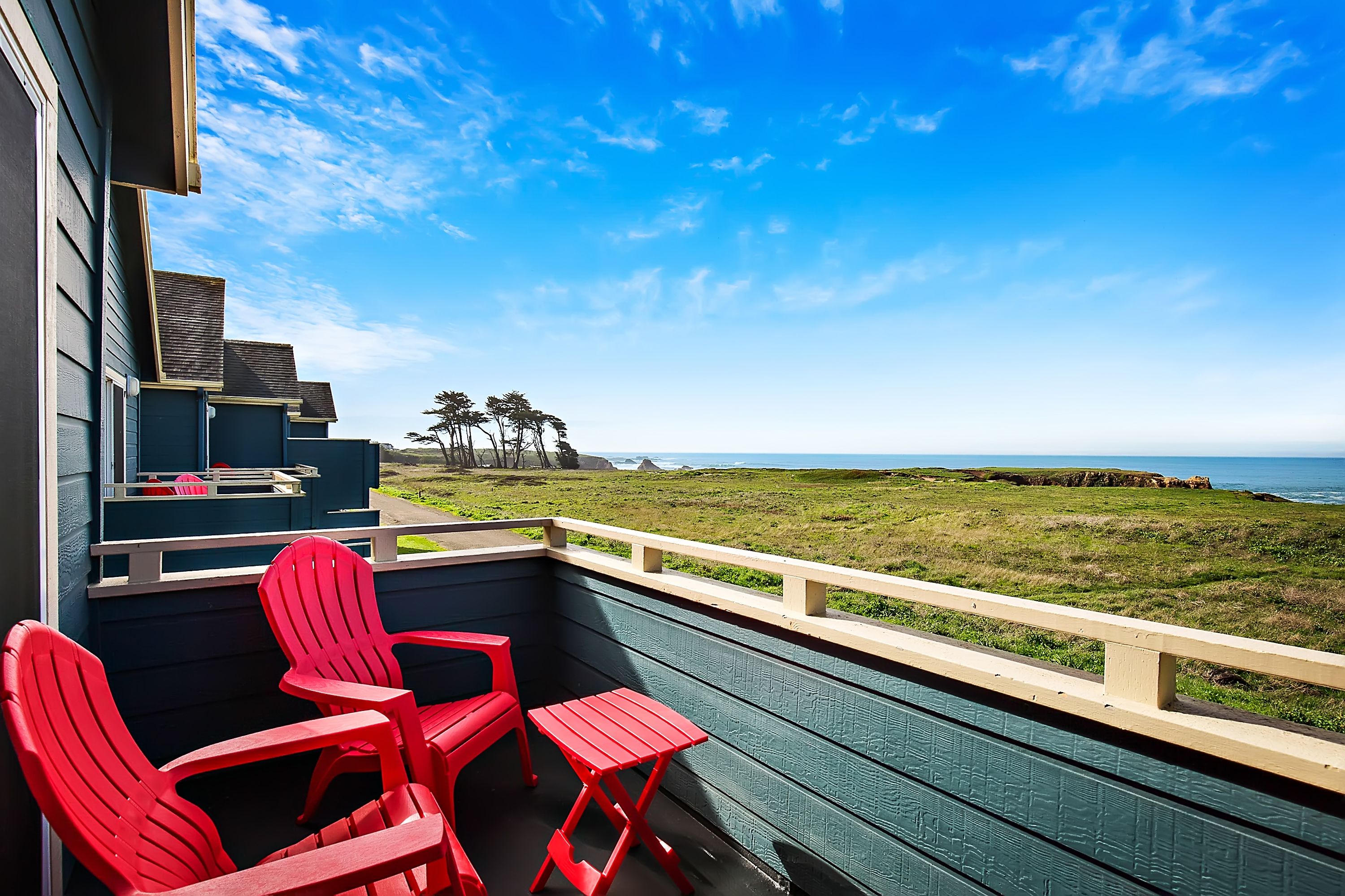 A balcony with red chairs and a small table, overlooking a grassy area and the ocean.