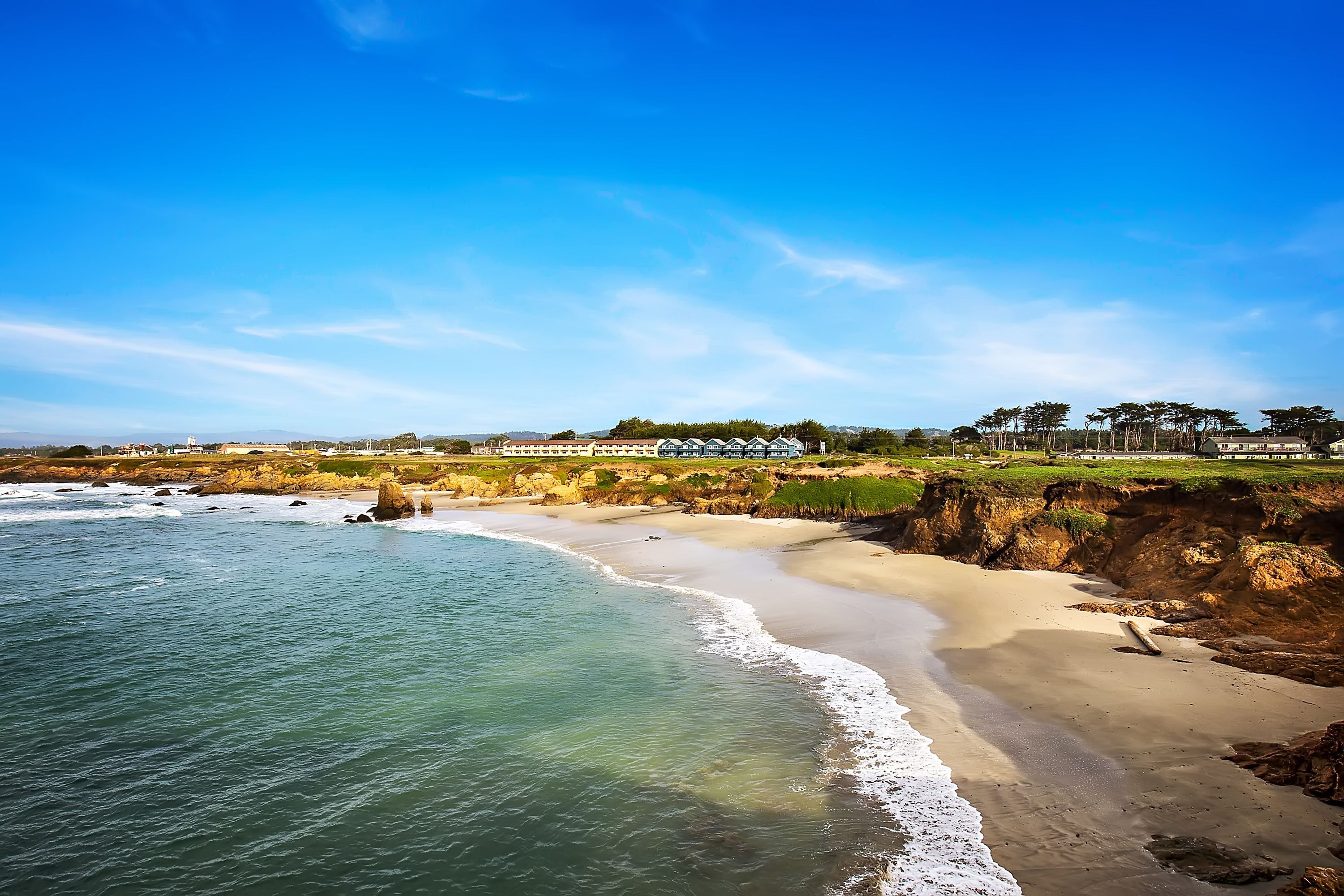 A coastal landscape with a sandy beach, ocean waves, and a row of buildings in the background.
