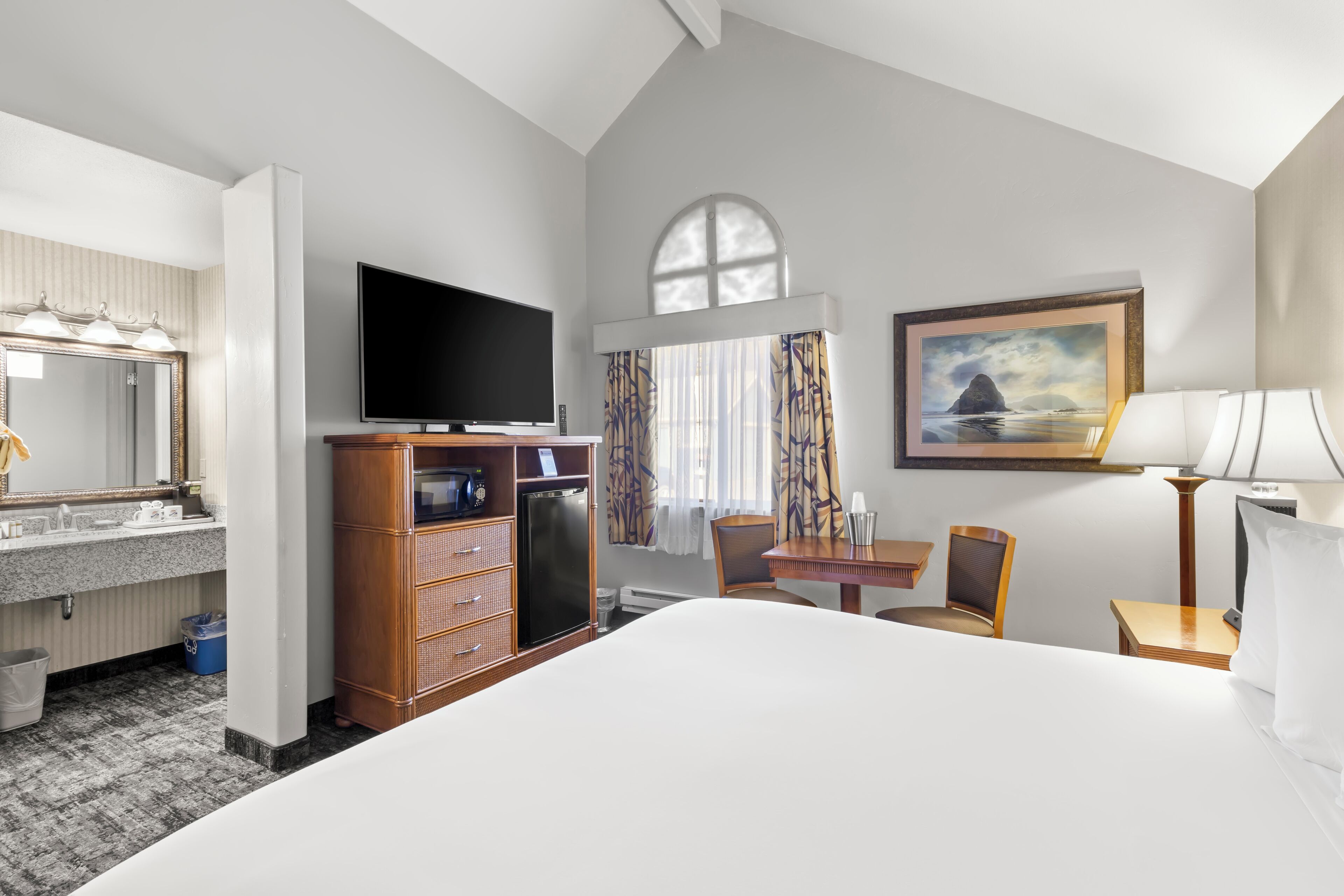 A hotel room with a large bed, a television, a desk with a chair, a small table, a lamp, a painting, a window with curtains, a mirror, a vanity with a sink, and a refrigerator.