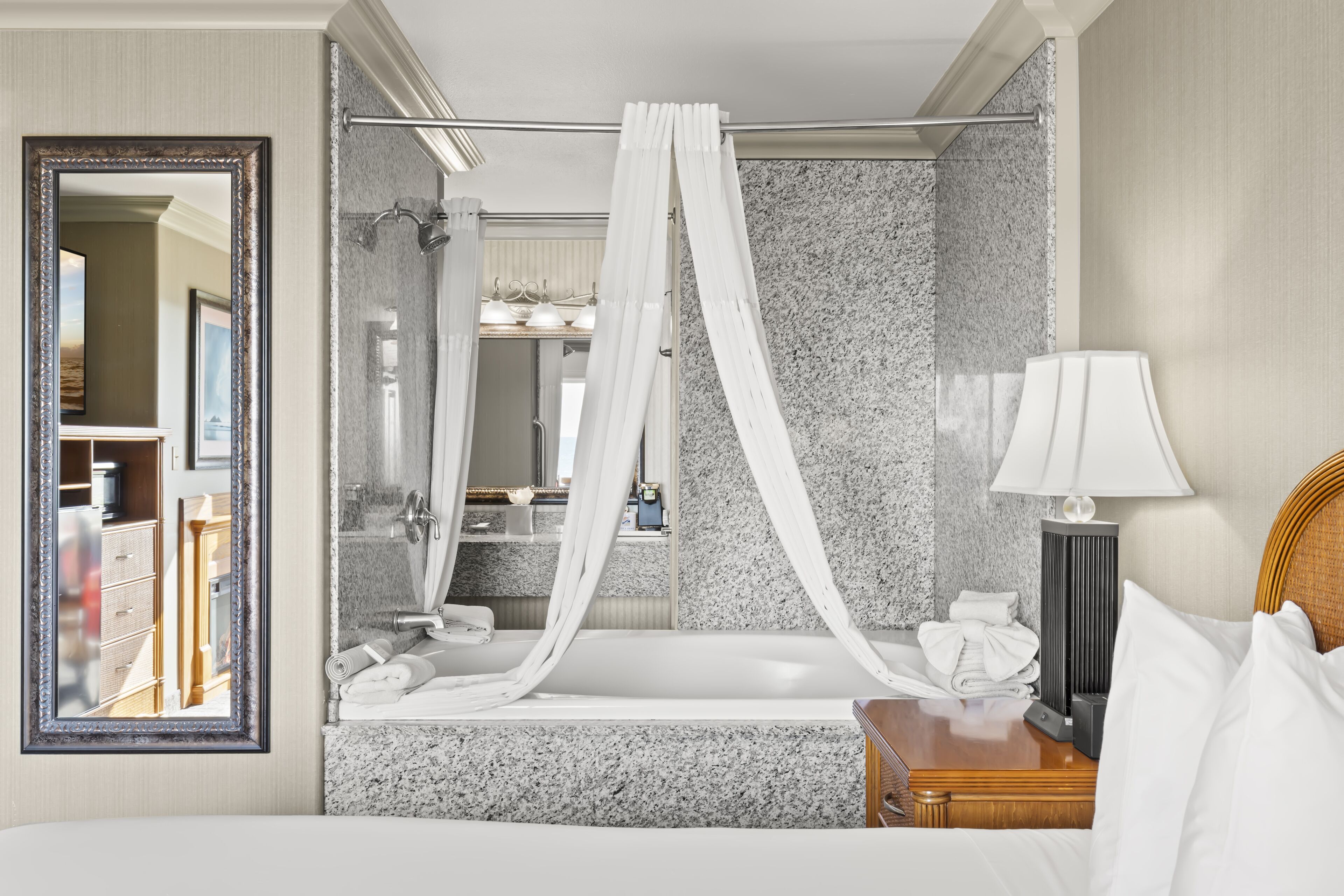 Ein Hotelzimmer mit einem großen Bett, einer Badewanne, einem Spiegel und einem Nachttisch mit einer Lampe.