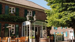 Een klassiek Europees gebouw met een bakstenen gevel, groene luiken en een overdekt terras met houten stoelen en tafels.