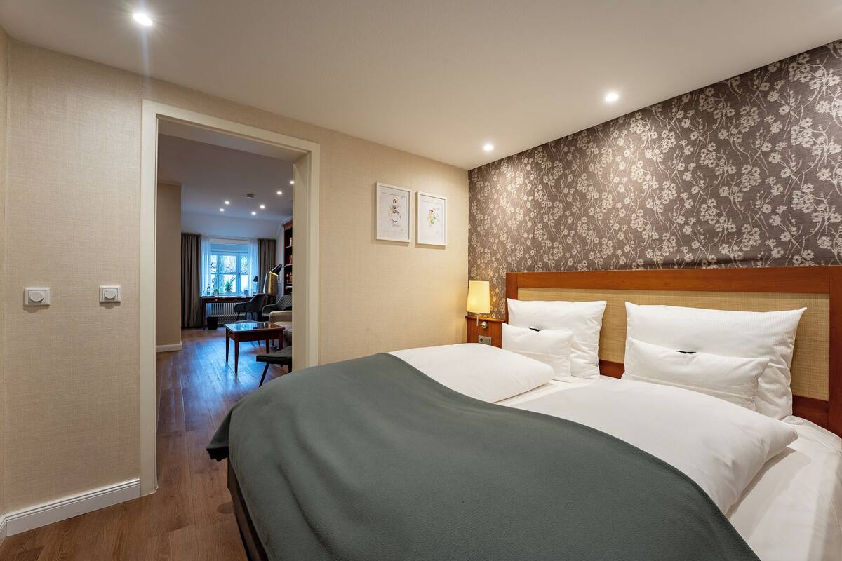A hotel room with a bed, a nightstand, a lamp, and a door leading to a hallway.