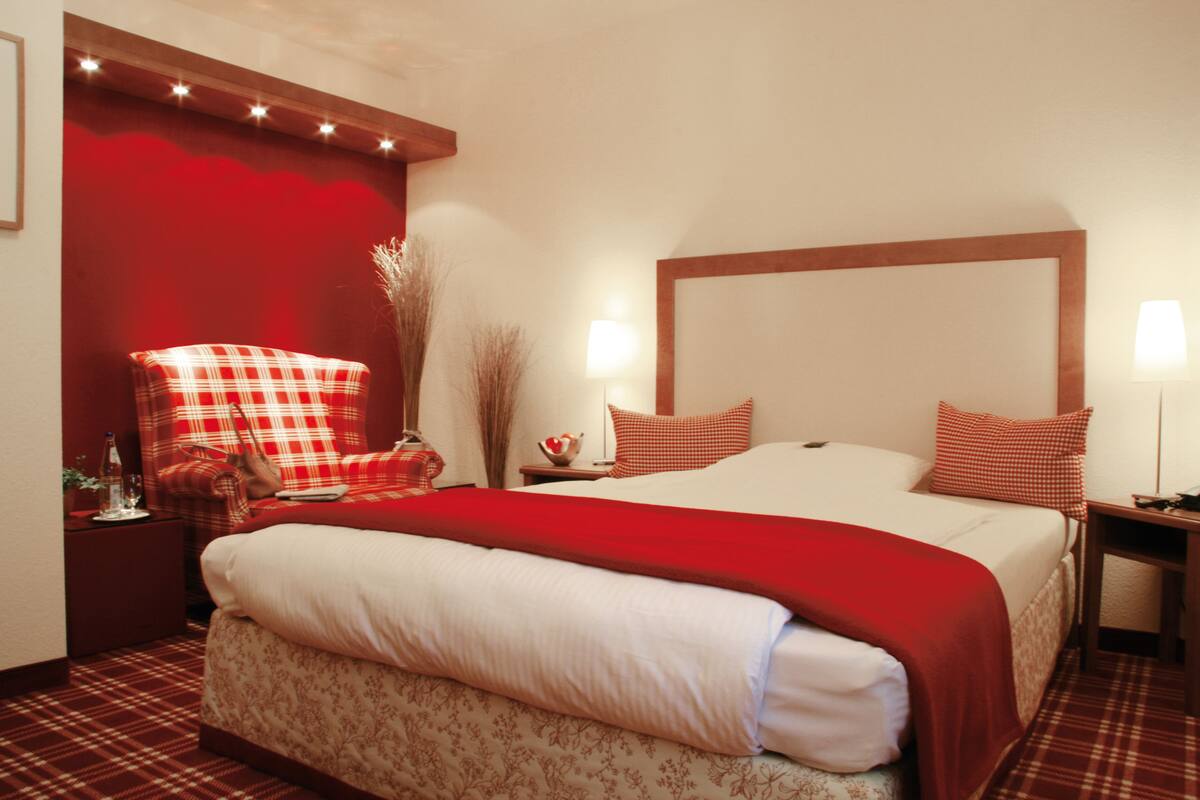 A hotel room with a large bed, a red and white checkered armchair, a bedside table with a lamp, and a red accent wall.