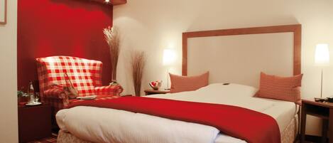 Una habitación de hotel con una cama grande, un sillón rojo y blanco a cuadros, una mesita de noche con una lámpara y una pared de acento roja.