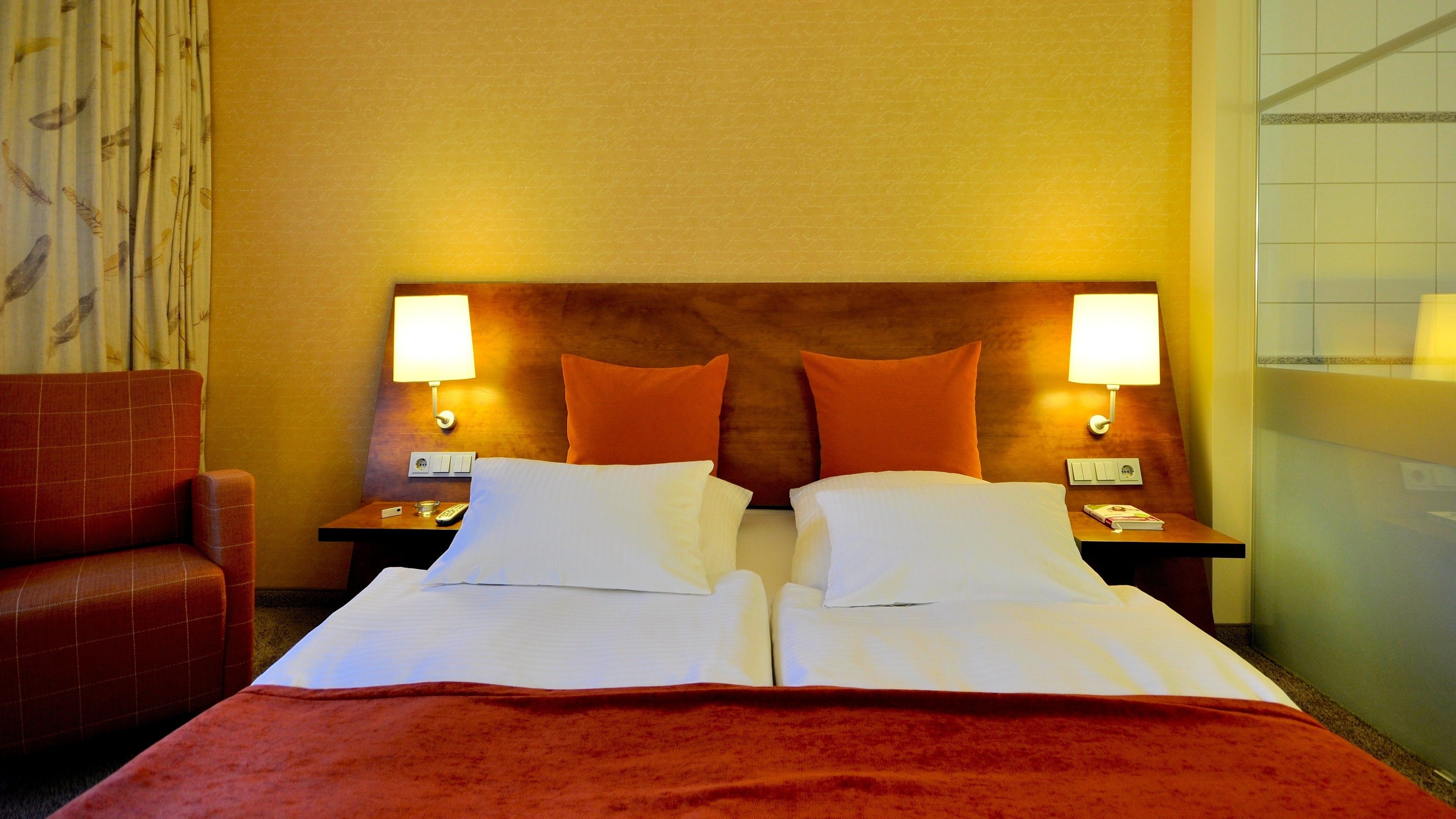 A double bed with white linens, two orange pillows, and a red blanket. There are two bedside tables with lamps, a red armchair, and a wall with a patterned curtain.