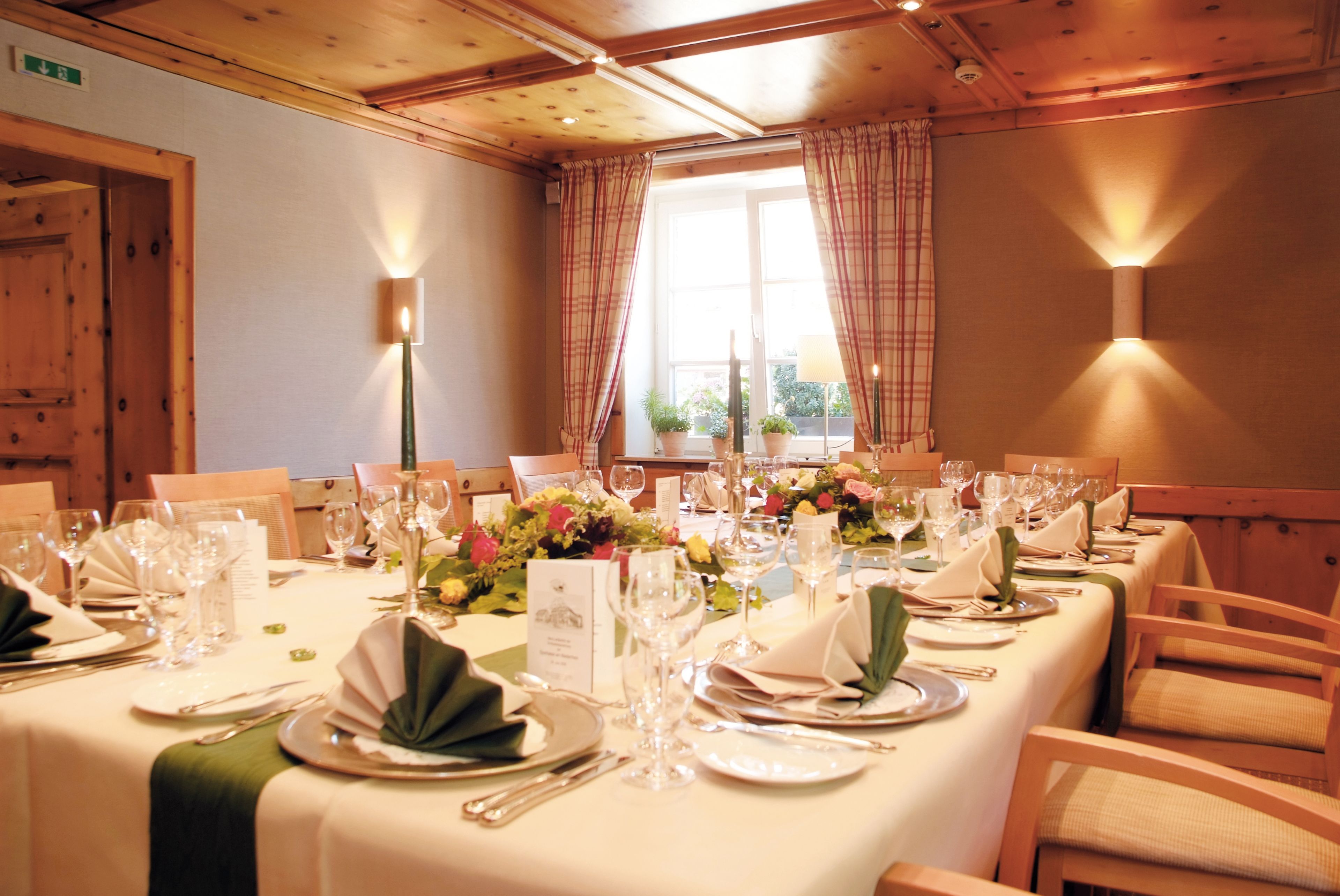 A dining room with a long table set for a meal, featuring multiple chairs, a window with curtains, and a wooden ceiling.