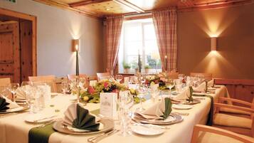 A dining room with a long table set for a meal, featuring multiple chairs, a window with curtains, and a wooden ceiling.