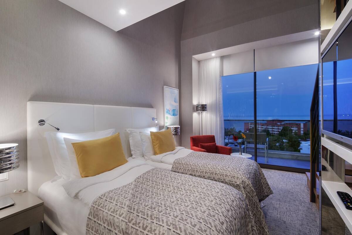 A modern hotel room with two beds, a red chair, a TV, and a balcony with a city view.