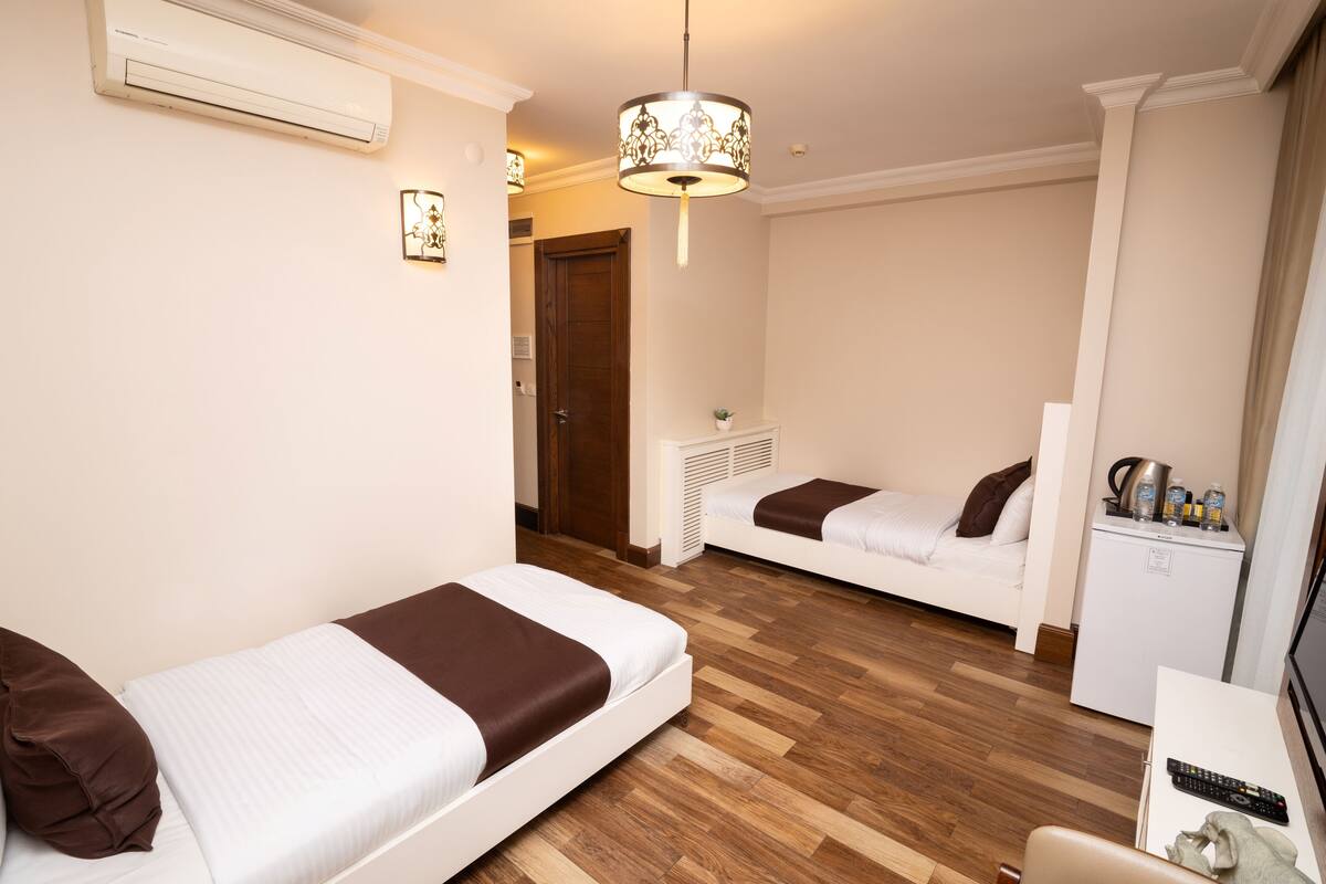 A hotel room with two beds, a desk with a chair, a small kitchenette, and a ceiling light fixture.