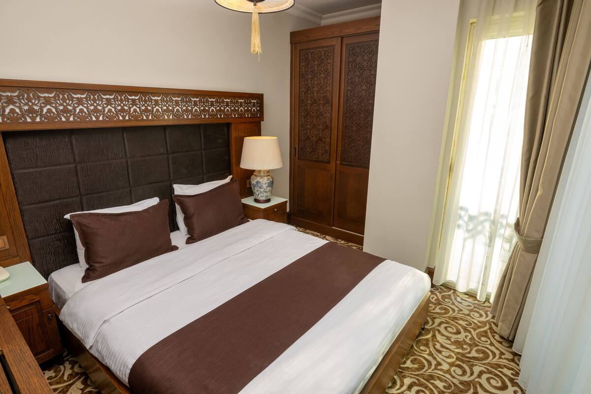 A hotel room with a large bed, a nightstand, a lamp, a decorative headboard, a wardrobe, and a window with curtains.