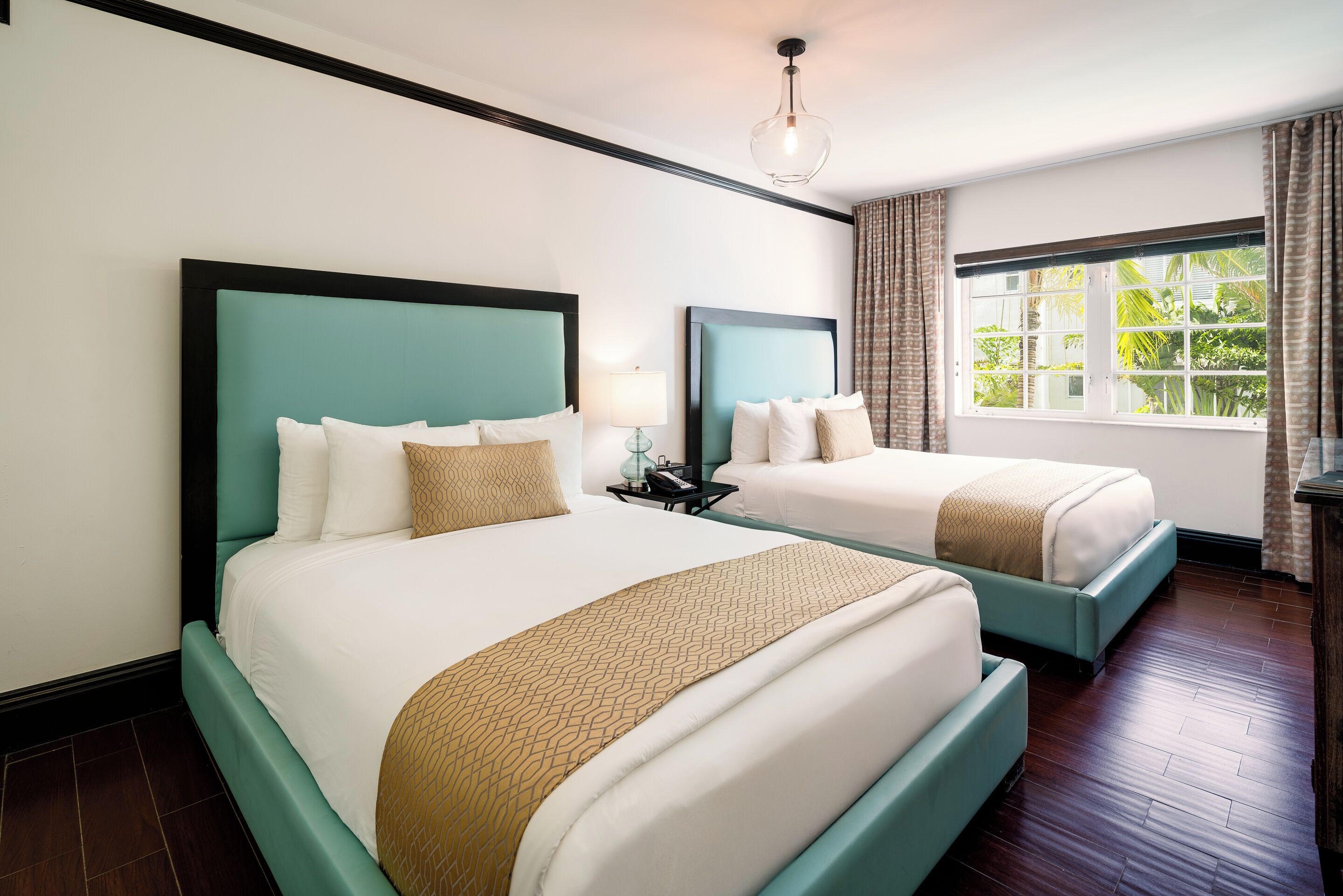 A hotel room with two beds, a nightstand, a lamp, and a window with curtains.