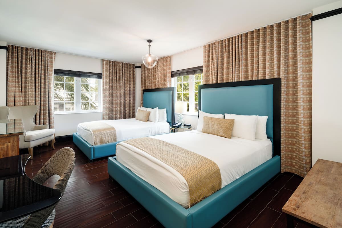 Ein Hotelzimmer mit zwei Betten, einem Schreibtisch, einem Stuhl und großen Fenstern mit Vorhängen.