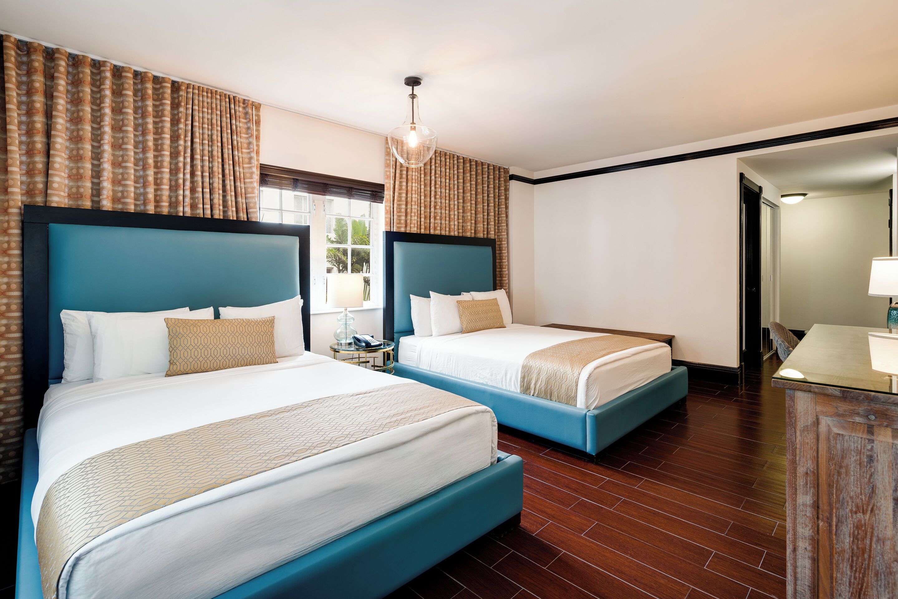 A hotel room with two beds, a wooden floor, a desk with a lamp, and a view through a window.