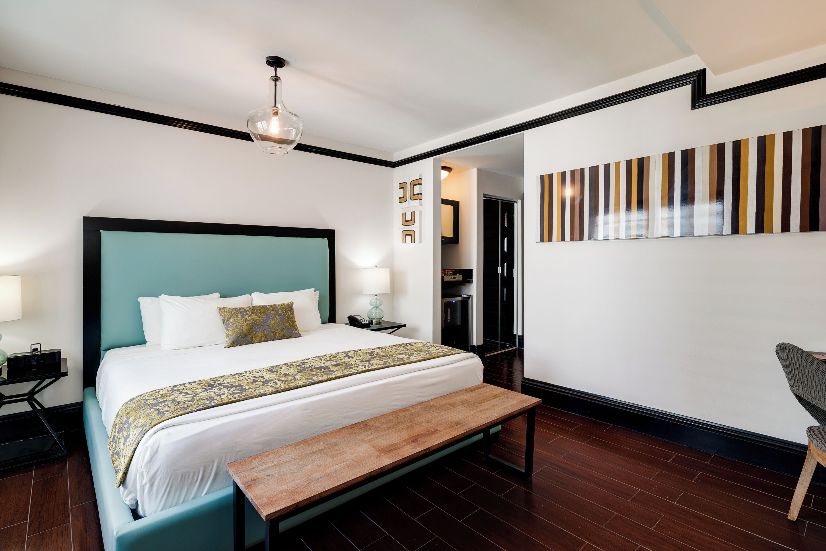 A modern hotel room with a large bed, a wooden bench, a desk, and a wall-mounted artwork.