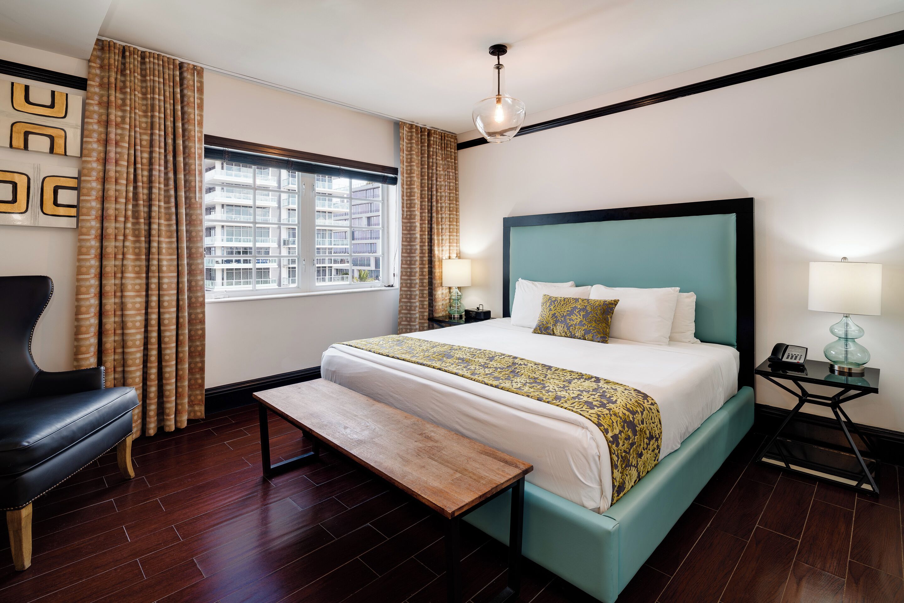 Een hotelkamer met een bed, een bankje, een stoel, een nachtkastje en een raam met gordijnen.